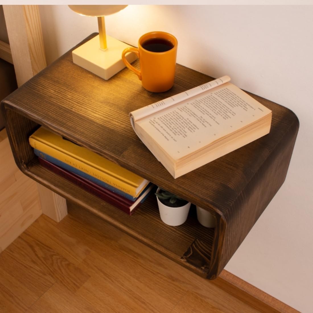 Curved wall-mounted solid wood floating nightstand with drawer and open shelf in light wood finish, styled with books and a bedside lamp